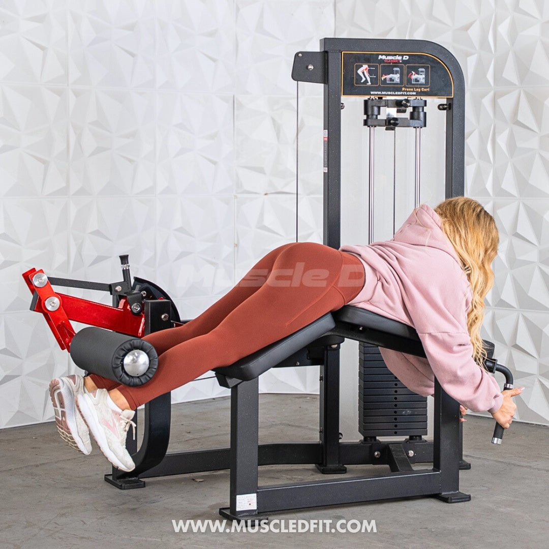 Person using a Classic-V2-Prone-Leg-Curl machine in a gym setting with a white textured wall background.