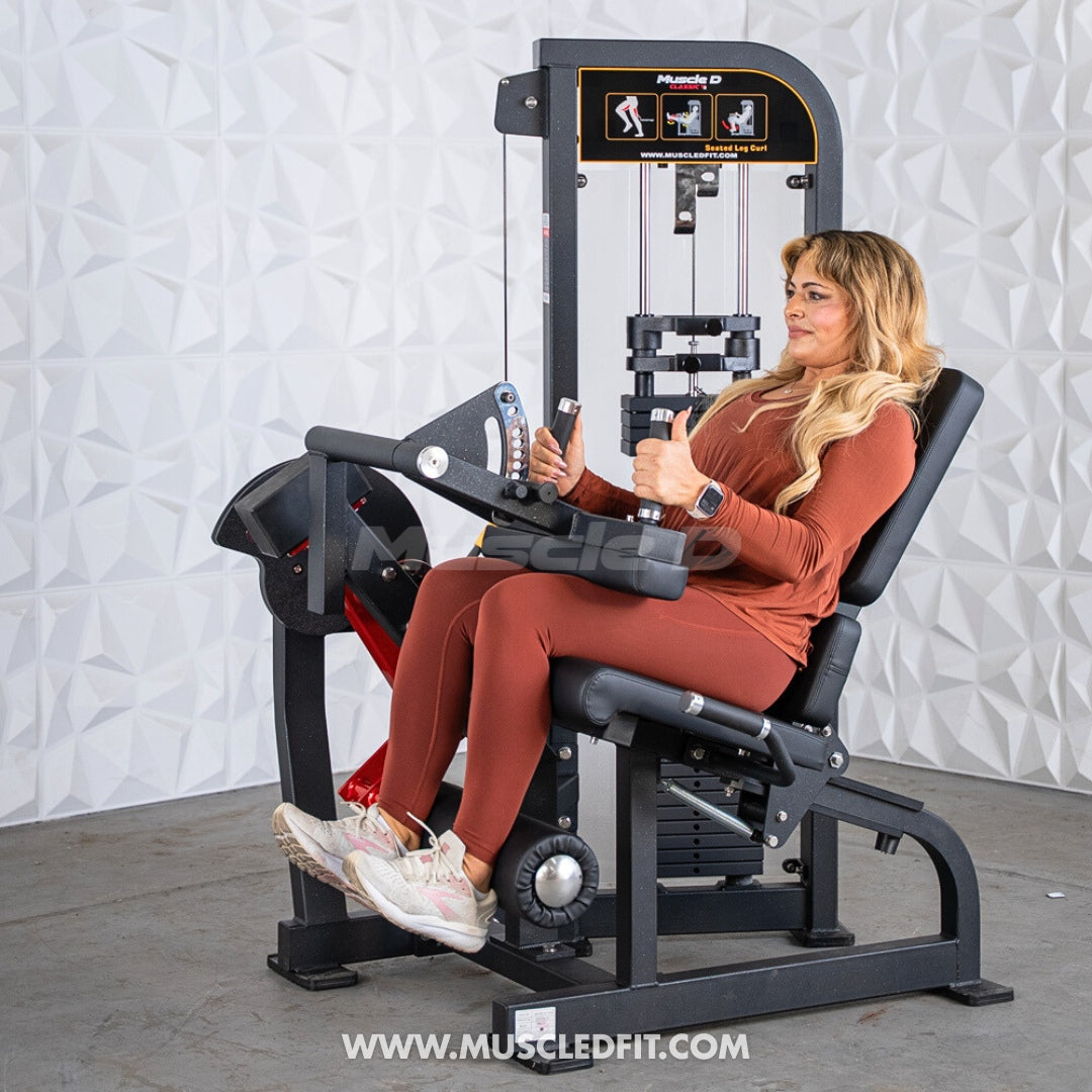 Woman using a Classic V2 Seated Leg Curl machine by Muscle D Fitness in a gym setting with a white textured wall background.