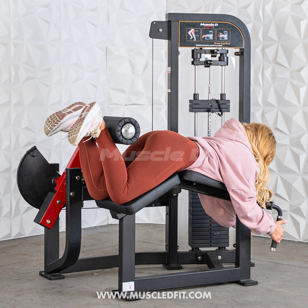 Person using a Classic V2 Prone Leg Curl machine by Muscle D Fitness against a textured wall.