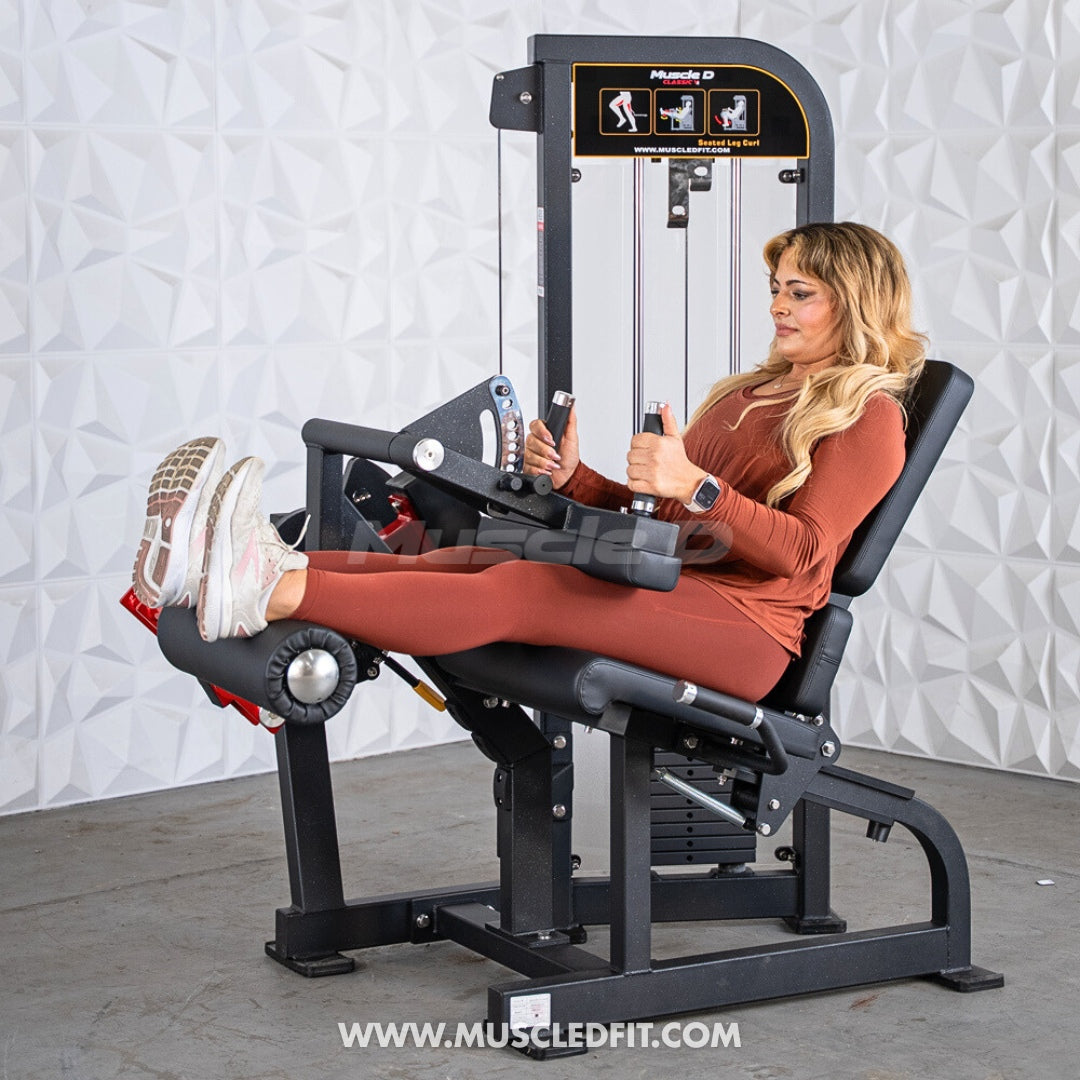 Woman using a Classic V2 Seated Leg Curl machine in a gym setting with a white textured wall background.